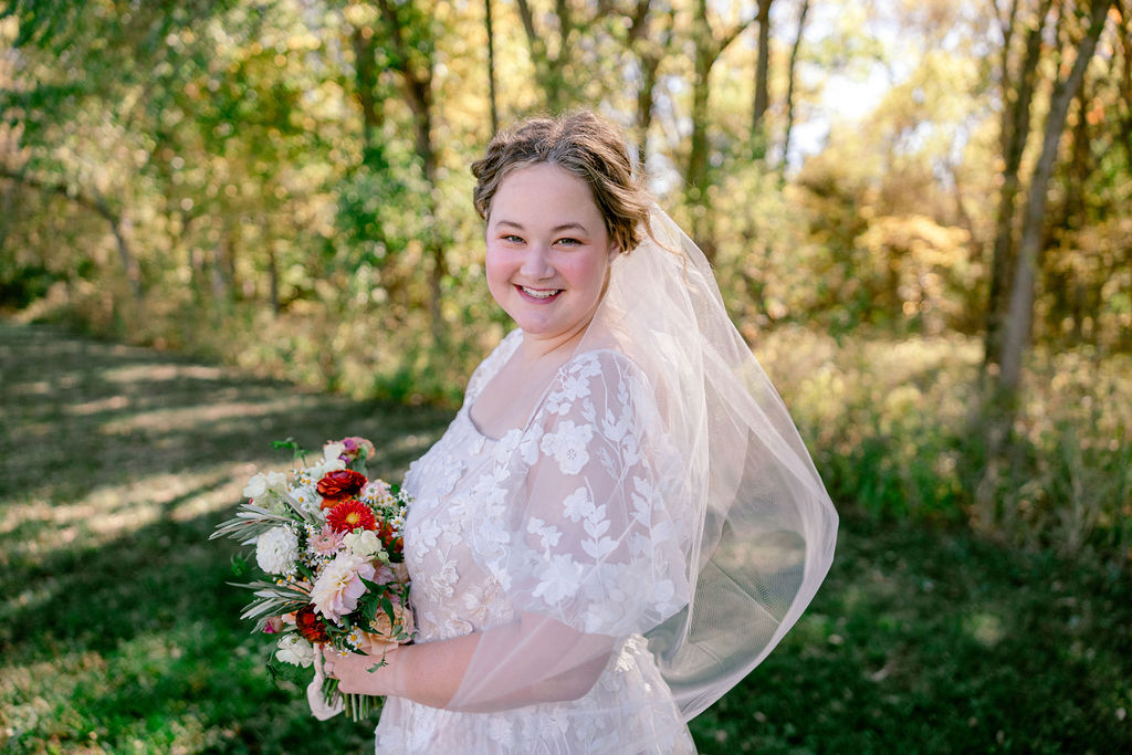 Bride smiles at camera with bouquet of rust, pink and white with forest backdrop.